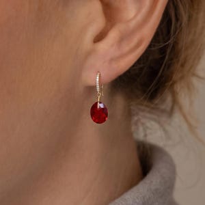 May include: Gold-toned hoop earrings with a small, clear stone-studded band and a faceted, oval-shaped, deep red gemstone pendant. The earrings are shown on a person's ear, with a neutral background.