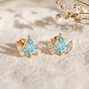 Könnte beinhalten: Ein Paar goldene Ohrstecker. Jeder Ohrring hat einen hellblauen, rautenförmigen Edelstein in der Mitte, umgeben von kleineren, klaren Edelsteinen. Die Ohrringe sind vor einem weichen, neutralen Hintergrund platziert.