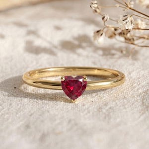 May include: A gold ring featuring a single heart-shaped red gemstone set in the center. The band is smooth and polished, showcasing a minimalist design that highlights the vibrant color and faceted cut of the central heart-shaped stone.