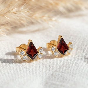 Puede incluir: Un par de pendientes de oro. Cada pendiente presenta una piedra de color rojo oscuro en forma de diamante en el centro, rodeada de piedras más pequeñas y transparentes. Los pendientes están colocados sobre un fondo de tela blanca, con plantas secas en el fondo.