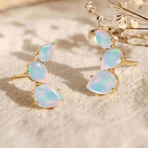 Könnte beinhalten: Ein Paar goldfarbene Ohrstecker, jeweils mit drei tropfenförmigen Opalen. Die Opale weisen einen schimmernden, irisierenden Effekt mit Blau-, Rosa- und Weißtönen auf. Die Ohrringe sind vor einem neutralen, strukturierten Hintergrund platziert.
