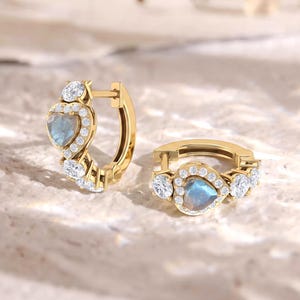 Peut inclure: Une paire de créoles en or avec une pierre bleue irisée en forme de cœur au centre, entourée d'un halo de petites pierres rondes transparentes. Les boucles d'oreilles présentent un mécanisme de fermeture à charnière et une finition dorée.