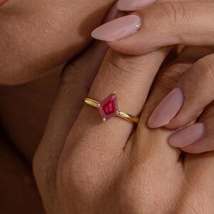 May include: A gold ring featuring a kite-shaped, deep pink gemstone set in a simple, polished gold band. The ring is worn on a finger, showcasing the geometric cut of the stone and the smooth, reflective quality of the gold metal setting.
