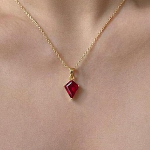 May include: A gold chain necklace featuring a kite-shaped pendant with a deep red gemstone. The pendant is suspended from a delicate gold link chain that rests against the collarbone, showcasing a polished metal finish and a faceted red stone.