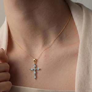 Puede incluir: Un collar dorado con un colgante de cruz adornado con piedras preciosas de color azul claro. El collar se lleva sobre una parte superior de color neutro.