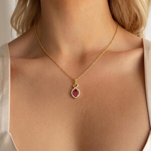 Può includere: Una collana a catena dorata con un ciondolo caratterizzato da una gemma ovale color rosa intenso al centro, circondata da un alone di piccole pietre chiare. Il design del ciondolo include un piccolo elemento a forma di cuore in alto.