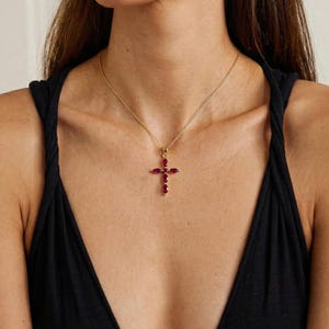 Könnte beinhalten: Eine goldfarbene Halskette mit einem Kreuzanhänger. Das Kreuz ist mit ovalen, tiefroten Edelsteinen besetzt. Die Halskette wird von einer Person getragen, die ein schwarzes Oberteil trägt.