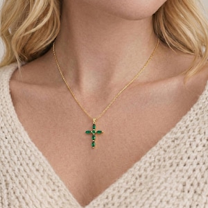 Puede incluir: Un collar dorado con un colgante en forma de cruz adornado con piedras preciosas verdes. La cruz cuelga en el centro del pecho. El fondo es un suéter color crema.