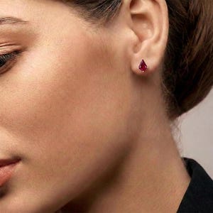 Pode incluir: Um close-up da orelha de uma pessoa com um brinco de botão em forma de pipa de cor rubi. O brinco é cravejado em metal dourado. O tom de pele da pessoa é claro e o fundo está desfocado.