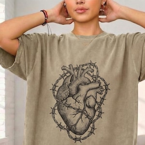 以下が含まれることがあります： オリーブグリーンのTシャツ。黒と白で描かれた、有刺鉄線に囲まれた人間の心臓のイラストが特徴です。半袖でゆったりとしたフィット感があり、普段使いに適しています。
