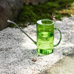 May include: A transparent green watering can with a curved handle and a long, silver spout. The can is filled with water and sits on a bed of small white stones. The watering can is designed for indoor or outdoor plant care.