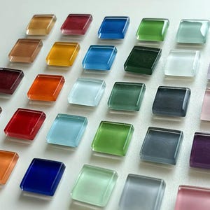 May include: A collection of colorful, square glass tiles in various shades, including amber, red, blue, green, and purple. The tiles are arranged in a grid pattern, showcasing their translucent quality and diverse hues. Ideal for mosaic art or decorative projects.