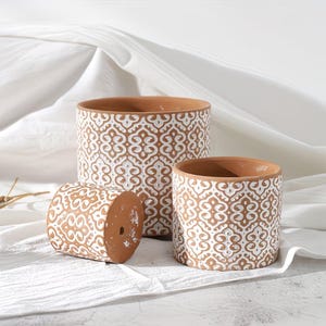 May include: Three terracotta-colored ceramic plant pots with a white, repeating geometric pattern. The pots are cylindrical, with one lying on its side. The largest pot is in the background, with a medium and small pot in front.