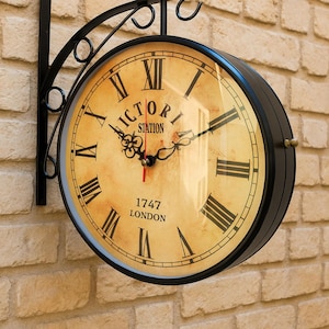 May include: A double-sided, black metal wall clock with a vintage design. The clock face is cream-colored with black Roman numerals and hands. The text "VICTORIA STATION 1747 LONDON" is printed on the face. The clock is mounted on a brick wall.