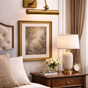May include: A brass wall-mounted picture light with an adjustable arm, positioned above a framed landscape painting. The painting features a forest scene in muted earth tones, enclosed in a gold-colored frame with a white mat board border.