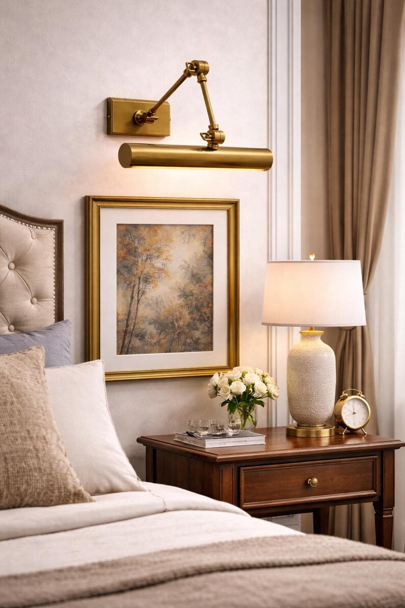 May include: A brass wall-mounted picture light with an adjustable arm, positioned above a framed landscape painting. The painting features a forest scene in muted earth tones, enclosed in a gold-colored frame with a white mat board border.