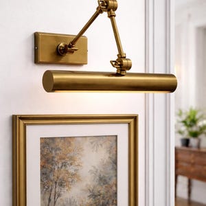 May include: A wall-mounted brass picture light with an adjustable swing arm and a rectangular backplate. The cylindrical light fixture features a metallic finish and is positioned above a framed artwork with a gold-toned frame on a white wall.