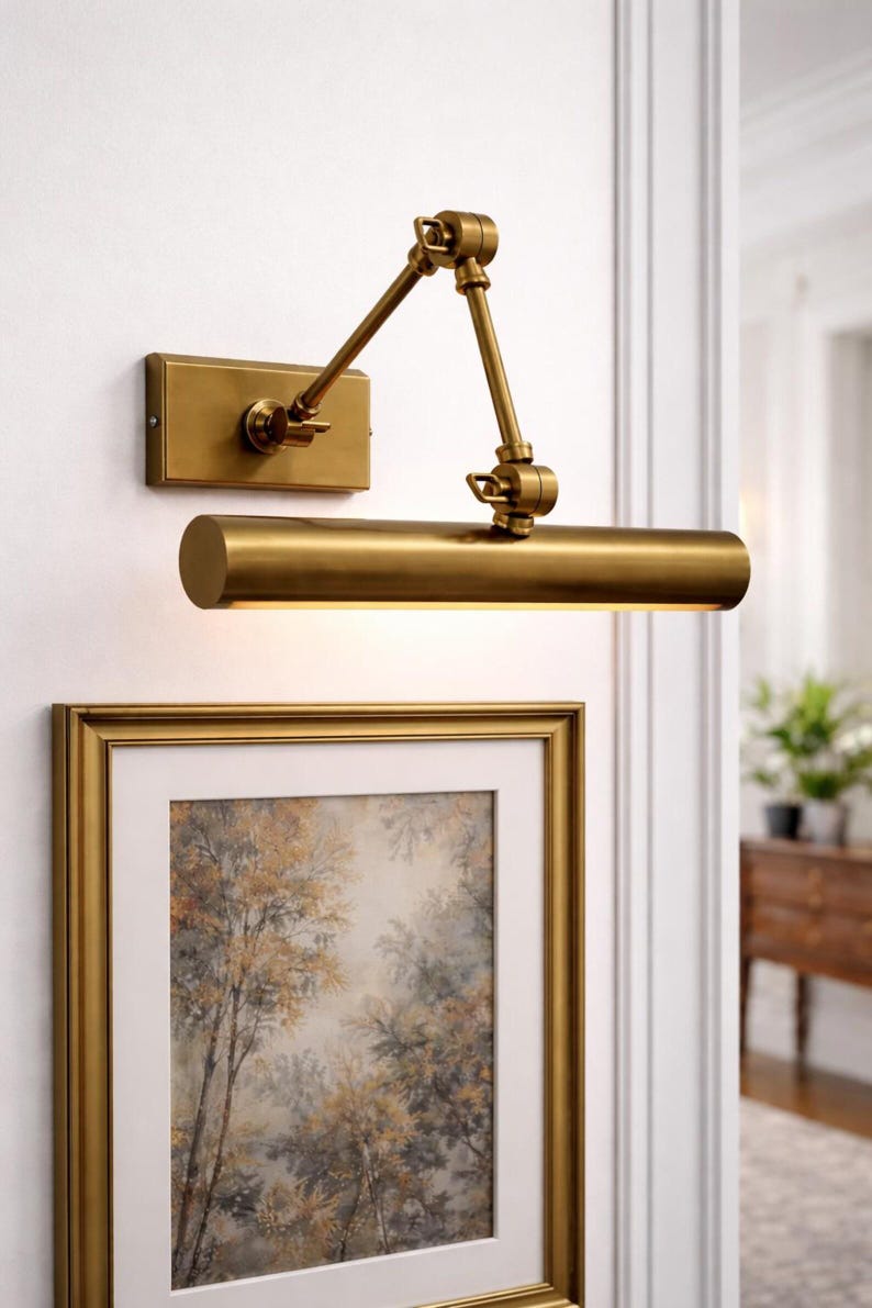 May include: A wall-mounted brass picture light with an adjustable swing arm and a rectangular backplate. The cylindrical light fixture features a metallic finish and is positioned above a framed artwork with a gold-toned frame on a white wall.