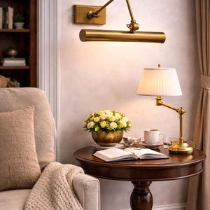May include: A brass wall-mounted picture light with an adjustable arm and a cylindrical shade, positioned above a round dark wood side table featuring a brass table lamp with a white pleated shade, a bowl of white roses, and an open book.