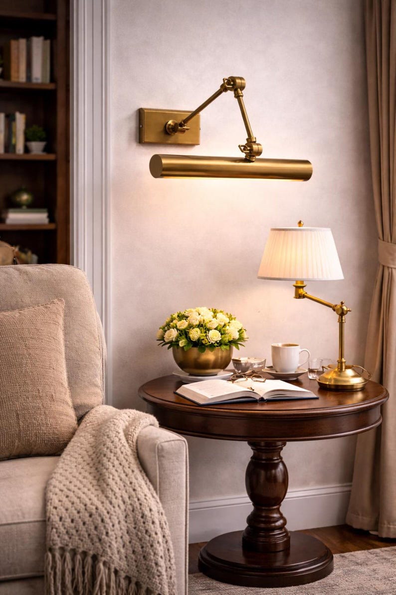 May include: A brass wall-mounted picture light with an adjustable arm and a cylindrical shade, positioned above a round dark wood side table featuring a brass table lamp with a white pleated shade, a bowl of white roses, and an open book.