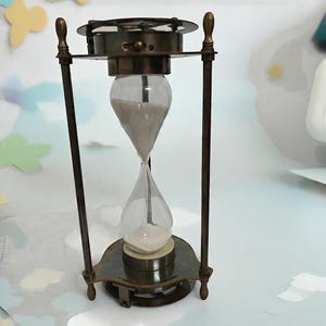 Könnte beinhalten: Eine antike Sanduhr mit Messingrahmen und klaren Glaskolben. Die Sanduhr enthält hellfarbenen Sand und wird von vier schlanken Messingstäben getragen. Ober- und Unterseite sind kreisförmig mit dekorativen Details.