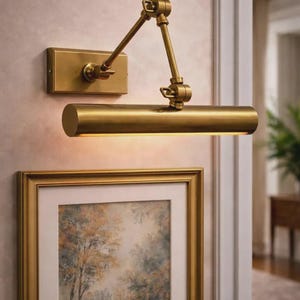 May include: A wall-mounted brass picture light with a rectangular backplate and an adjustable two-arm swing mechanism. The cylindrical light fixture features a warm metallic finish and is positioned directly above a framed artwork on the wall.