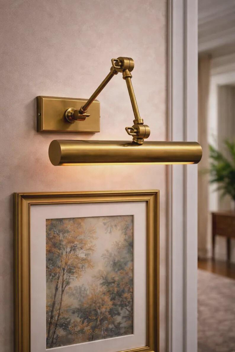 May include: A wall-mounted brass picture light with a rectangular backplate and an adjustable two-arm swing mechanism. The cylindrical light fixture features a warm metallic finish and is positioned directly above a framed artwork on the wall.
