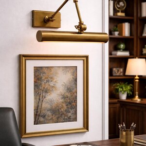 May include: A brass wall-mounted picture light with an adjustable swing arm and a cylindrical lamp head, positioned above a square framed painting of a forest scene with a white mat, featuring a metallic finish and a rectangular wall plate.