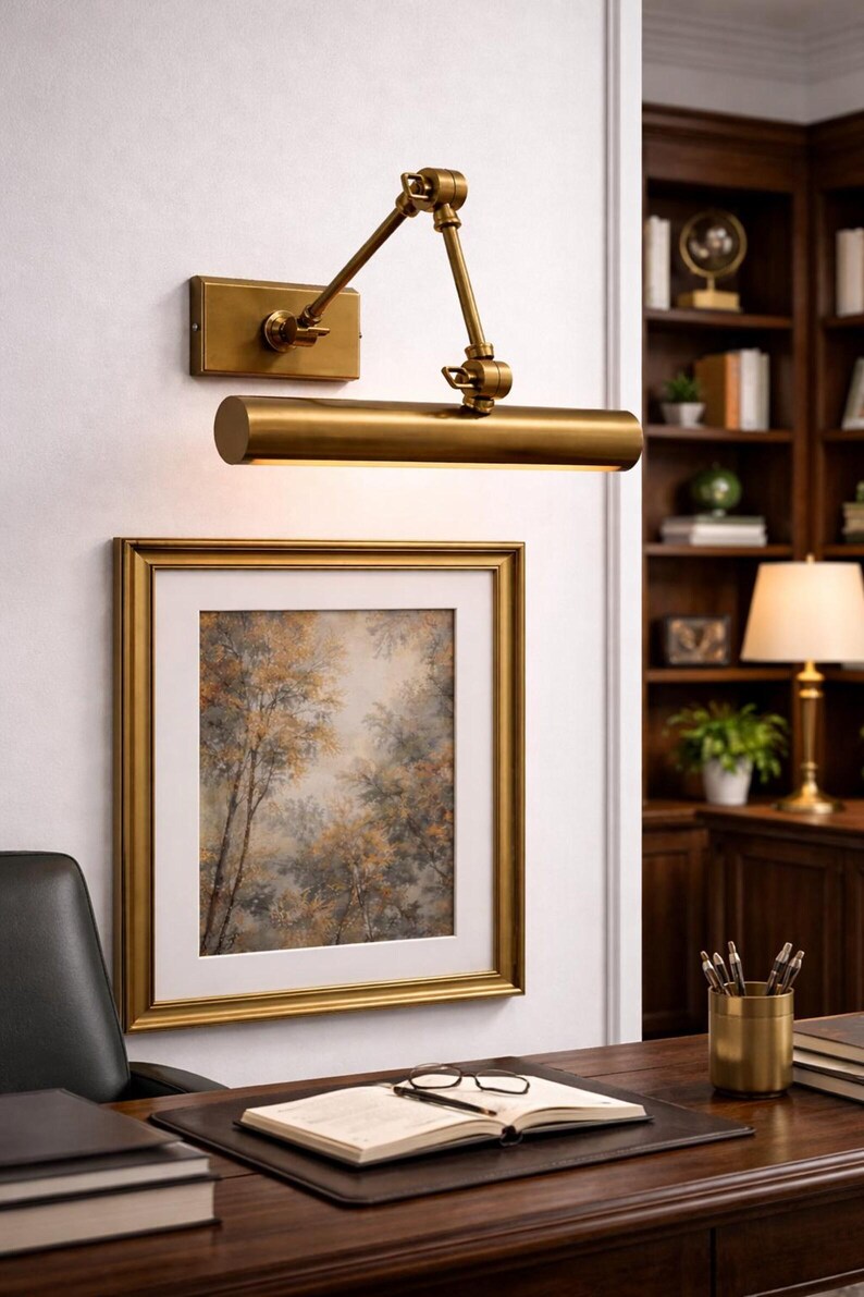 May include: A brass wall-mounted picture light with an adjustable swing arm and a cylindrical lamp head, positioned above a square framed painting of a forest scene with a white mat, featuring a metallic finish and a rectangular wall plate.