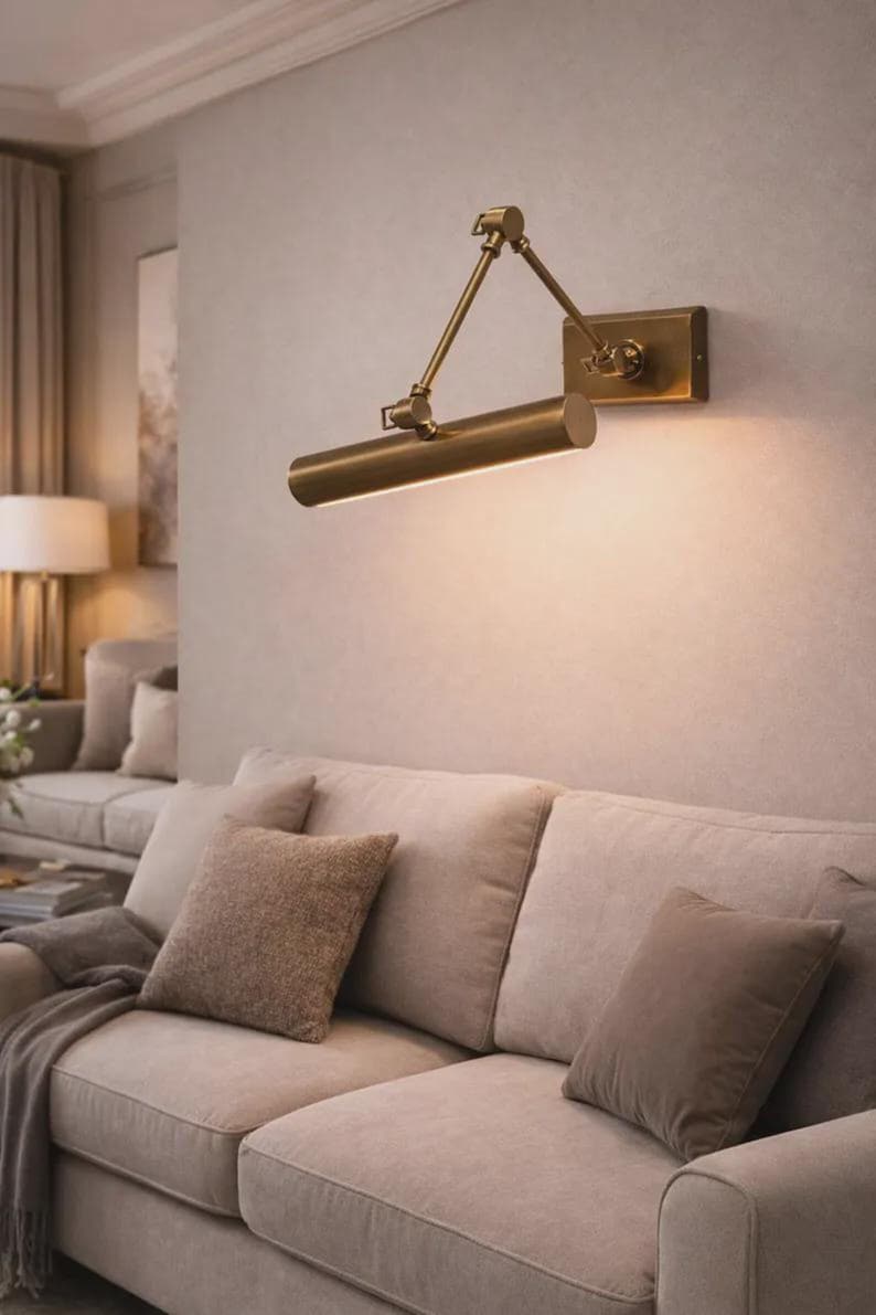 May include: A wall-mounted picture light with a metallic brass finish, featuring an adjustable swing arm and a cylindrical lamp head that emits a warm downward glow. The fixture is attached to a rectangular base plate mounted on the wall.