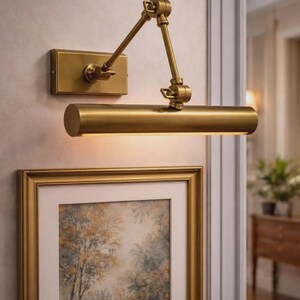 May include: A wall-mounted picture light with a brushed brass finish, featuring a rectangular base plate, an adjustable double-jointed arm, and a cylindrical light housing that directs illumination downward onto a framed artwork below.