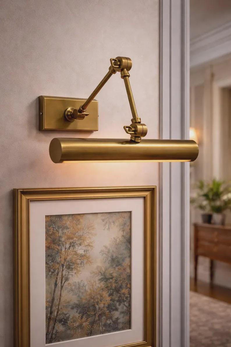 May include: A wall-mounted picture light with a brushed brass finish, featuring a rectangular base plate, an adjustable double-jointed arm, and a cylindrical light housing that directs illumination downward onto a framed artwork below.
