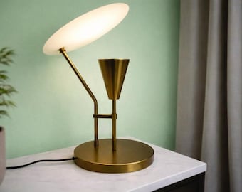 Peut inclure: Une lampe de table en laiton avec une base circulaire, une tige verticale et un élément en forme de cône. Un bras incliné soutient un abat-jour en verre blanc plat et ovale qui émet une lueur douce. La lampe possède un cordon noir.