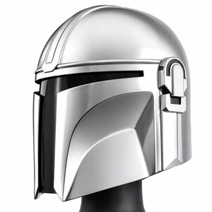 May include: A polished silver-colored helmet featuring a smooth, rounded dome, a T-shaped visor opening with a dark interior, and a prominent vertical ridge running along the center. The side includes a mechanical earpiece detail with geometric accents.