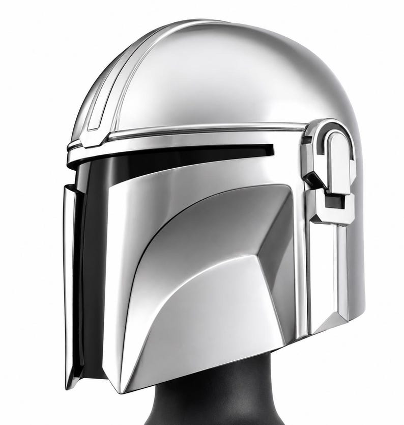 May include: A polished silver-colored helmet featuring a smooth, rounded dome, a T-shaped visor opening with a dark interior, and a prominent vertical ridge running along the center. The side includes a mechanical earpiece detail with geometric accents.