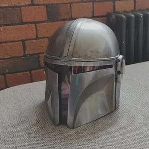 May include: A silver-colored helmet with a smooth, metallic finish and a T-shaped visor opening. The helmet features a rounded dome top, a central ridge running from front to back, and a distinct angular faceplate design with a dark, recessed visor area.