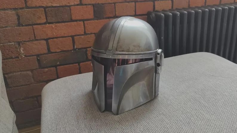 May include: A silver-colored helmet with a smooth, metallic finish and a T-shaped visor opening. The helmet features a rounded dome top, a central ridge running from front to back, and a distinct angular faceplate design with a dark, recessed visor area.