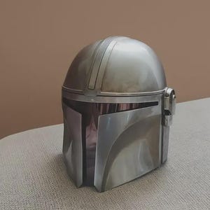 May include: A metallic silver helmet with a smooth, rounded dome and a T-shaped visor opening. The helmet features a central vertical ridge, a dark tinted face shield, and circular ear-piece details on the sides, finished with a polished sheen.