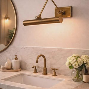 May include: A brass wall-mounted picture light with an adjustable swing arm, featuring a cylindrical lamp head and a rectangular base plate, positioned above a bathroom vanity with a matching brass faucet and two cross-handle controls.