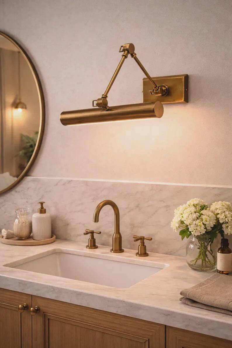 May include: A brass wall-mounted picture light with an adjustable swing arm, featuring a cylindrical lamp head and a rectangular base plate, positioned above a bathroom vanity with a matching brass faucet and two cross-handle controls.