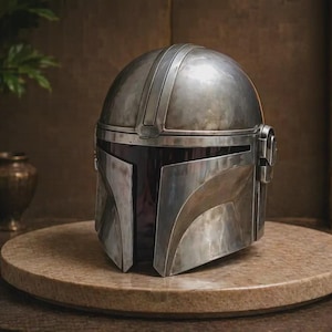 May include: A metallic silver helmet with a smooth, rounded dome and a T-shaped visor opening. The helmet features a weathered, brushed finish with subtle dark gray shading, a central ridge along the top, and circular side ear-piece details.