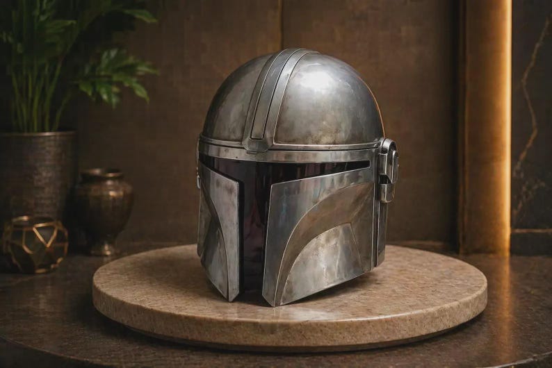 May include: A metallic silver helmet with a smooth, rounded dome and a T-shaped visor opening. The helmet features a weathered, brushed finish with subtle dark gray shading, a central ridge along the top, and circular side ear-piece details.