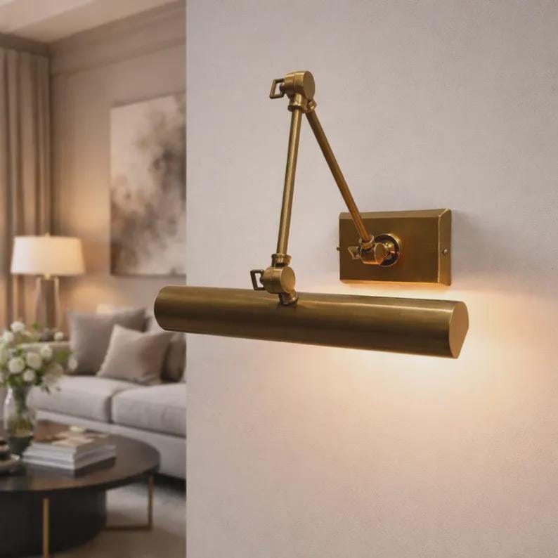 May include: A wall-mounted picture light with a brushed brass finish, featuring a rectangular backplate, an adjustable double-jointed arm, and a cylindrical light housing that directs illumination downward to highlight artwork or wall displays.