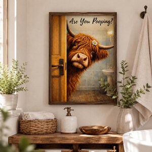Puede incluir: Un cuadro rectangular con marco de madera oscura que muestra una vaca Highland marr&oacute;n asom&aacute;ndose por una puerta de madera hacia un ba&ntilde;o alicatado. El texto Are You Pooping? aparece en letra cursiva negra sobre la vaca.
