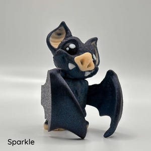 A small, dark blue vampire bat figurine with a sparkly glittery finish. The bat features large expressive eyes and cute pointy fangs.