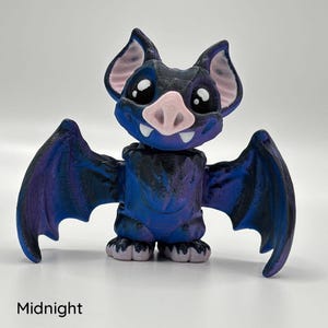 A 3D-printed Midnight coloured vampire bat fidget in shades of blue, black and purple, with little fangs. The bat has large, expressive eyes and is posed with its wings spread.