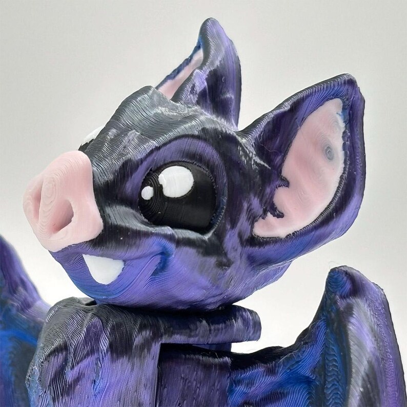 A 3D-printed bat figurine with a cartoon design. The bat has a purple and black body, large white eyes with black pupils, pink ears and nose, and two white fangs.
