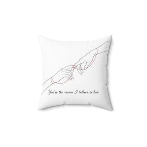 Può includere: Cuscino quadrato bianco con un design minimalista di due mani collegate da un filo rosso. Il testo "You're the reason I believe in love" è stampato in corsivo nero sotto.