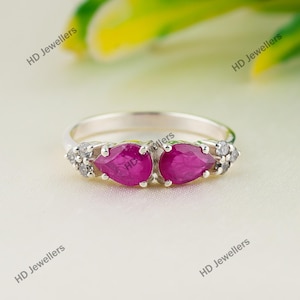 May include: A silver ring featuring two pear-cut, deep pink rubies set side-by-side in the centre, flanked by small, clear, round-cut accent stones on each side. The band is polished silver, and the text HD Jewellers appears on the image.