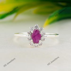 Puede incluir: Un anillo de plata con una piedra preciosa central ovalada de color rosa intenso rodeada por un halo de doce pequeñas piedras redondas transparentes. La banda es fina y de plata pulida. El texto HD Jewellers es visible.
